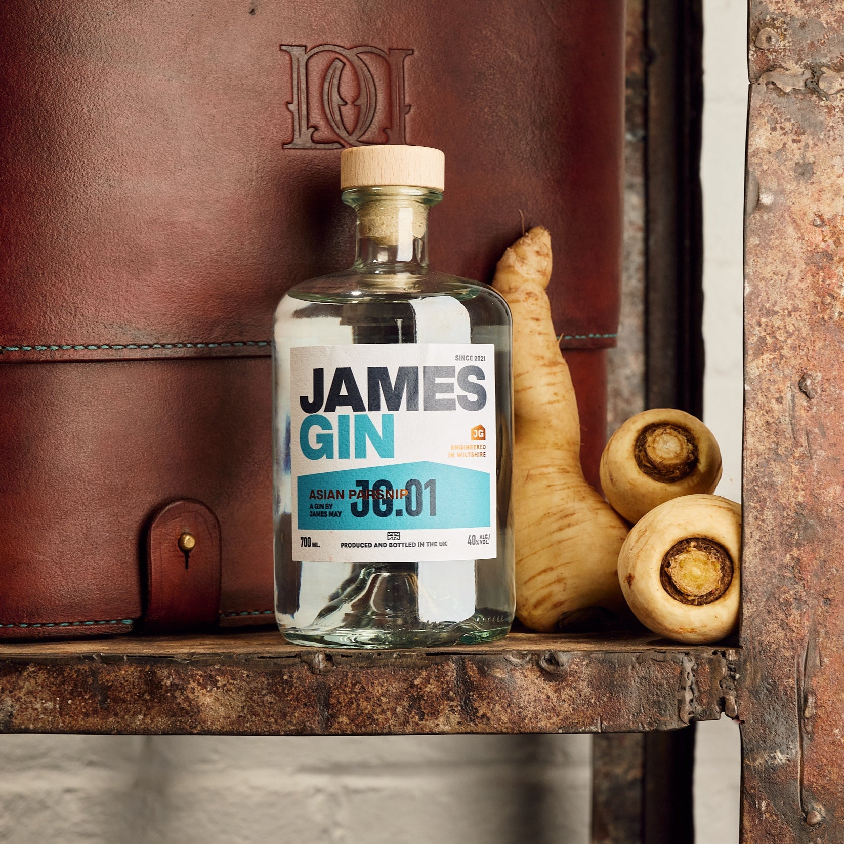 Bottle of James Gin Asian Parsnip on a rustic shelf beside some parsnips and a leather bag with the distillery logo on it