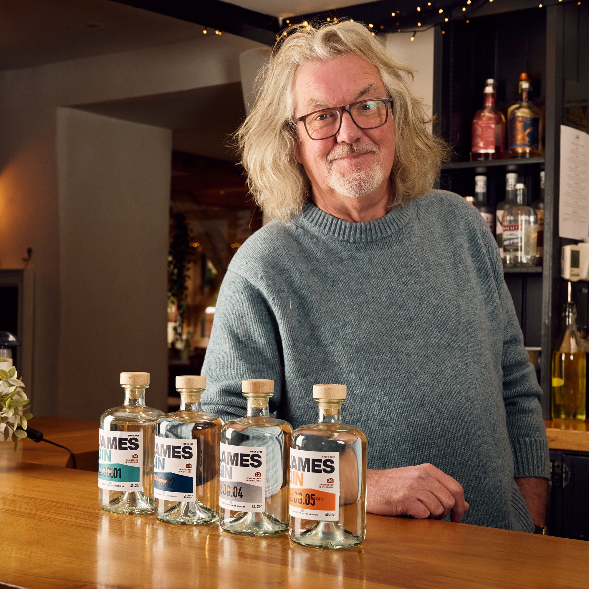 James May in his pub leaning on the bar alongside his range of four gins