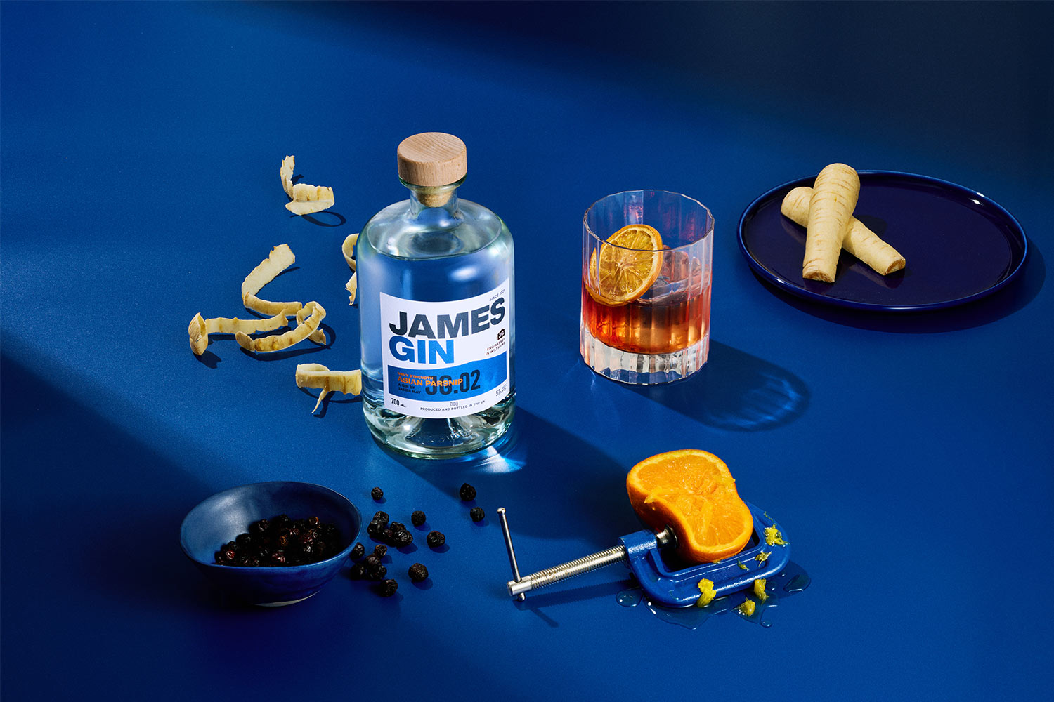 Bottle of James gin Asian Parsnip navy strength beside a negroni cocktail surrounded by parsnips, parsnip peel, small bowl of juniper berries and half an orange being squeezed by a vice.