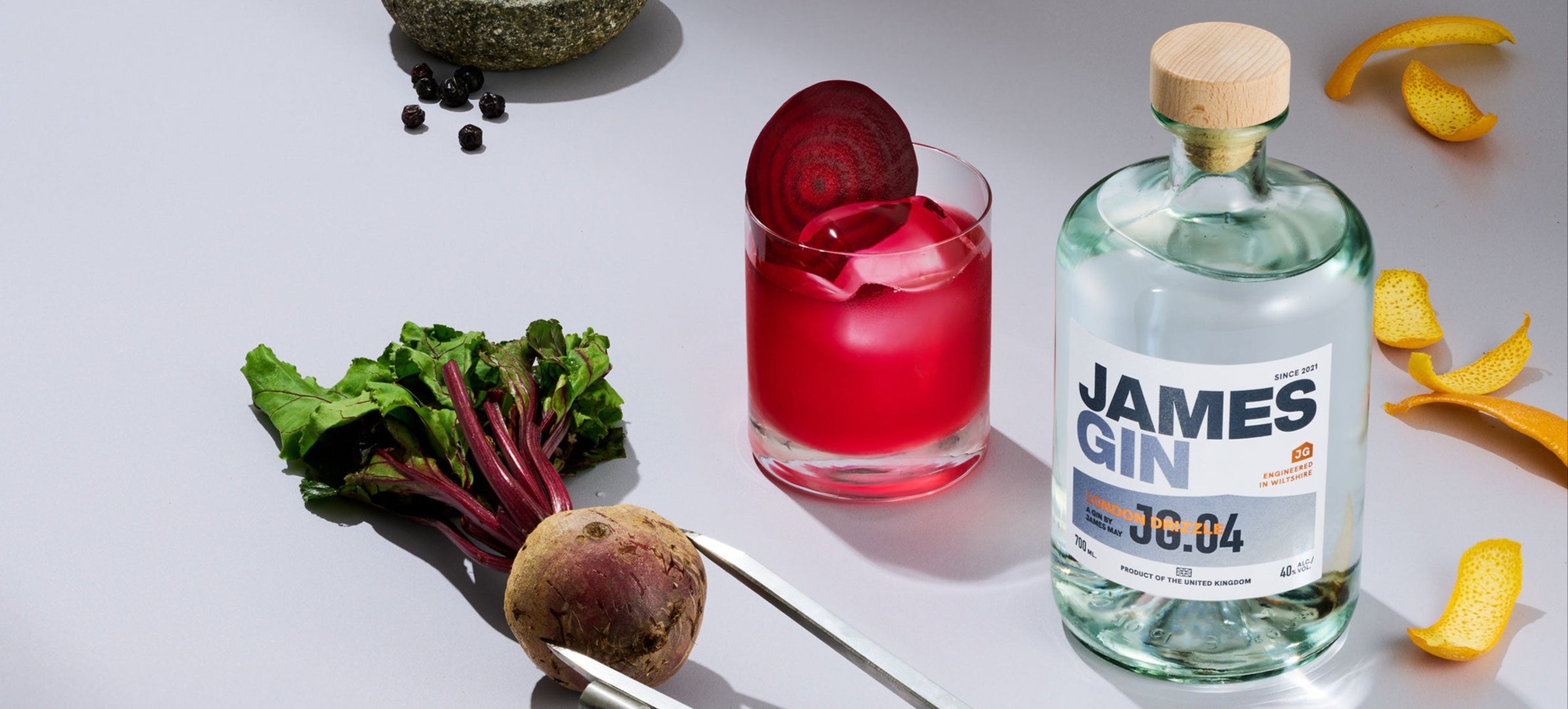 Bottle of James Gin London drizzle beside a beetroot cocktail, orange peel, a concrete bowl of juniper berries, and a compass measuring a beetroot
