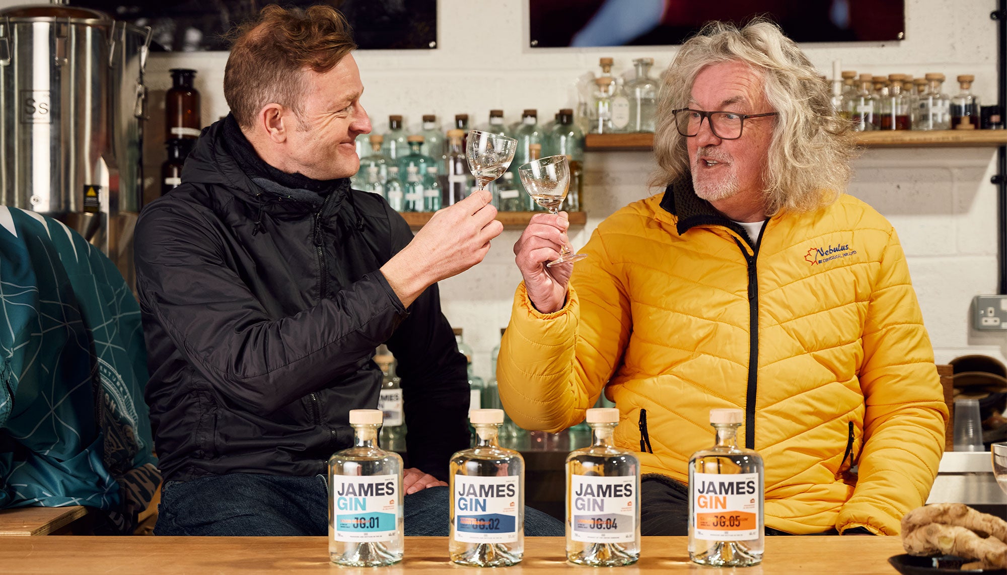 James and Hugh in the shed cheersing with glasses of gin behind a line up of four gin bottles