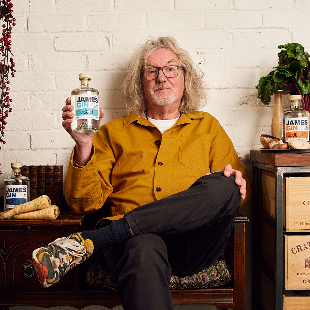 James May holding a bottle of his Asian Parsnip Gin, surrounded by other James Gin flavours and fresh ingredients.