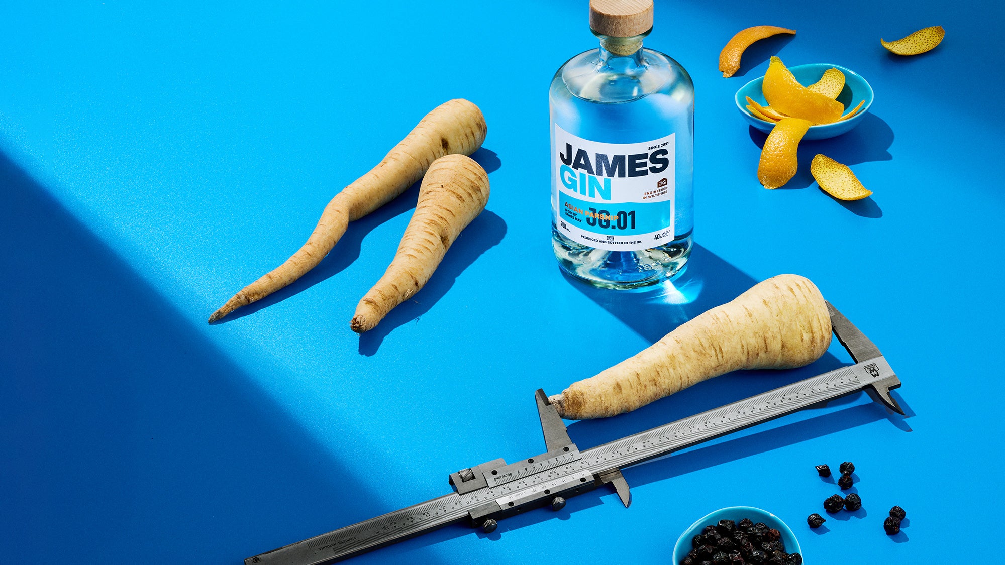 James Gin Asian Parsnip bottle developed by James May, with a parsnip being measured with callipers, dried juniper berries and orange peel in small bowls on a blue background.