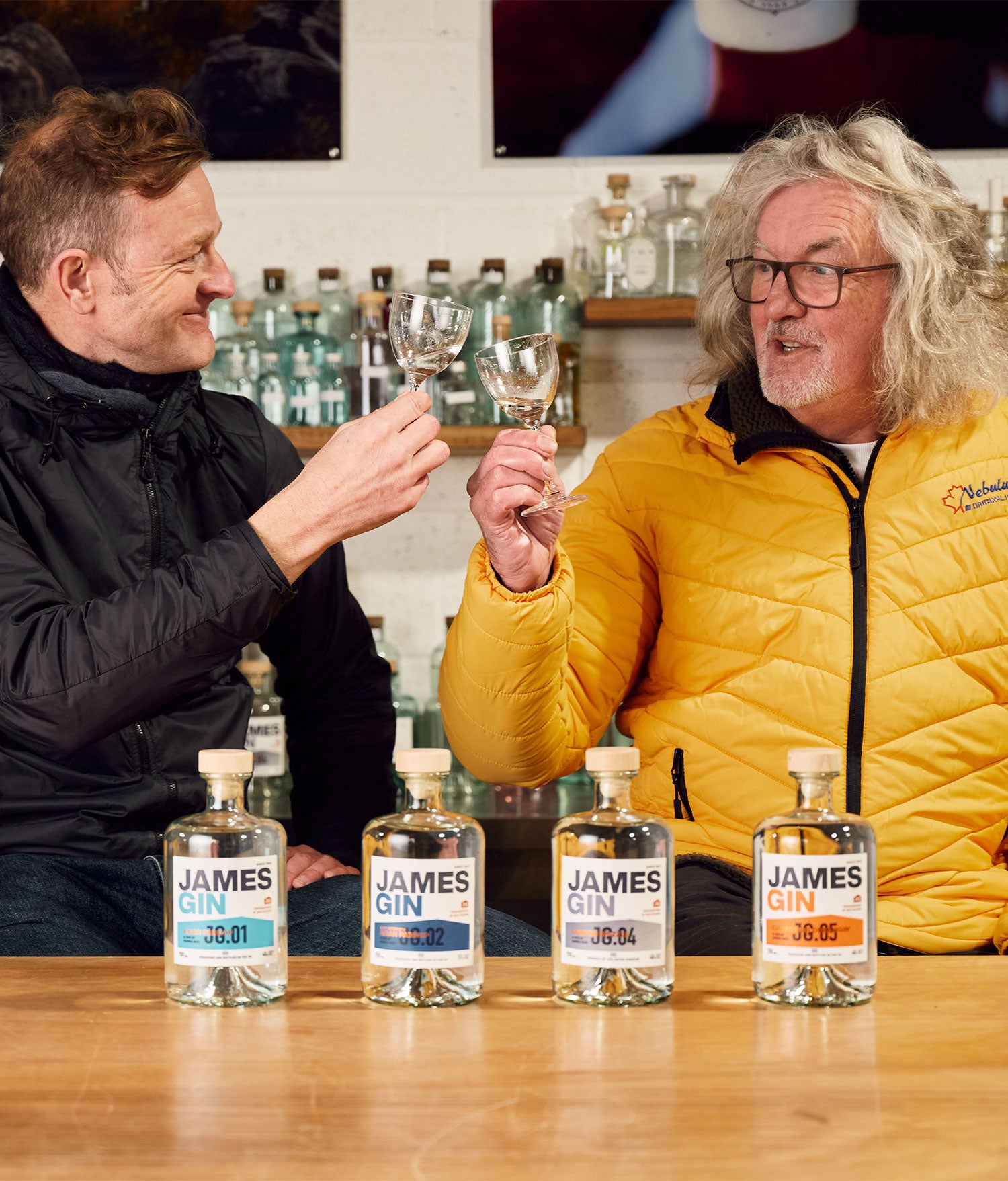 James and Hugh in the shed cheersing with glasses of gin behind a line up of four gin bottles