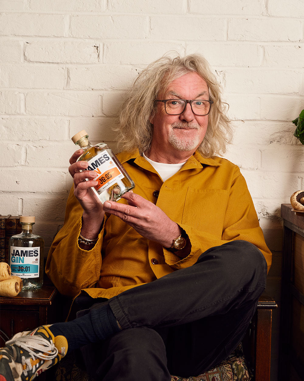 James May sitting in his workshop with his legs crossed holding a bottle of california dreamgin' beside his other gin bottles and parsnips