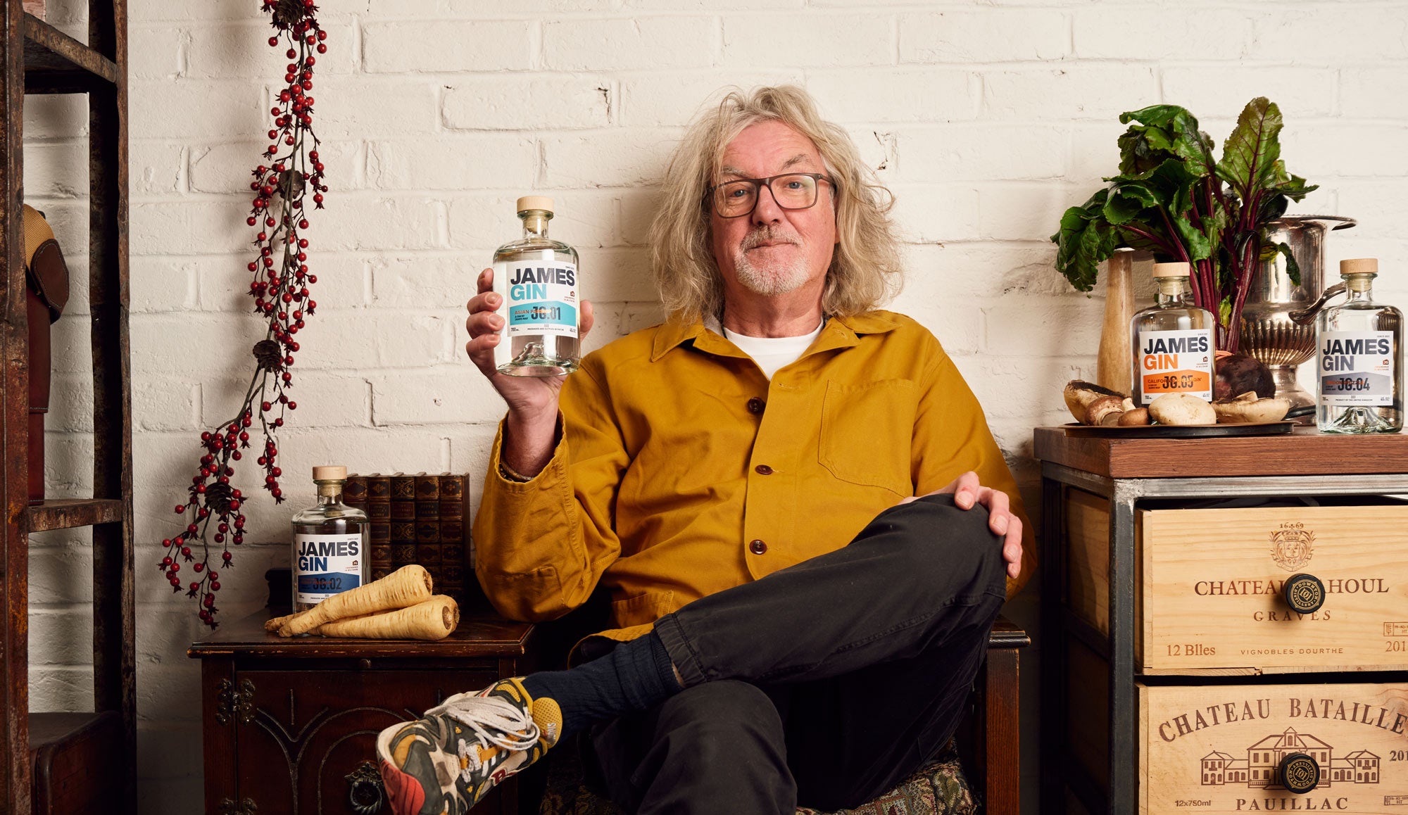 James May sitting in his workshop with his legs crossed, holding his asian parsnip gin surrounded by his other gin and botanicals such as parsnips and mushrooms