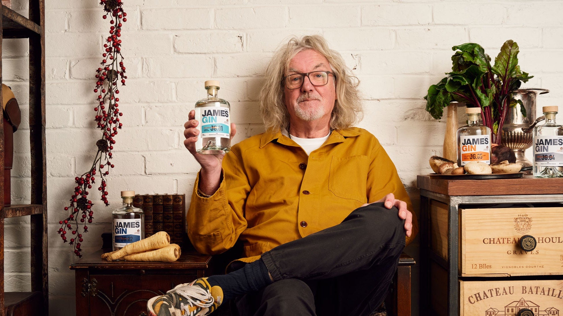James May holding a bottle of his Asian Parsnip Gin, surrounded by other James Gin flavours and fresh ingredients.