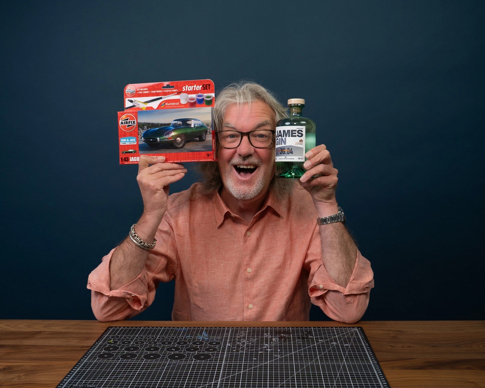 James May holding a bottle of James Gin and a model car kit against a dark blue background