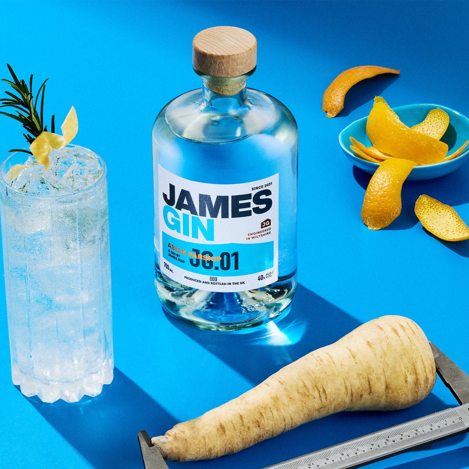 A James gin bottle of asian parsnip alongside a  cocktail. a parsnip being measured by a callipers, a bowl of juniper berries and a bowl of orange peel