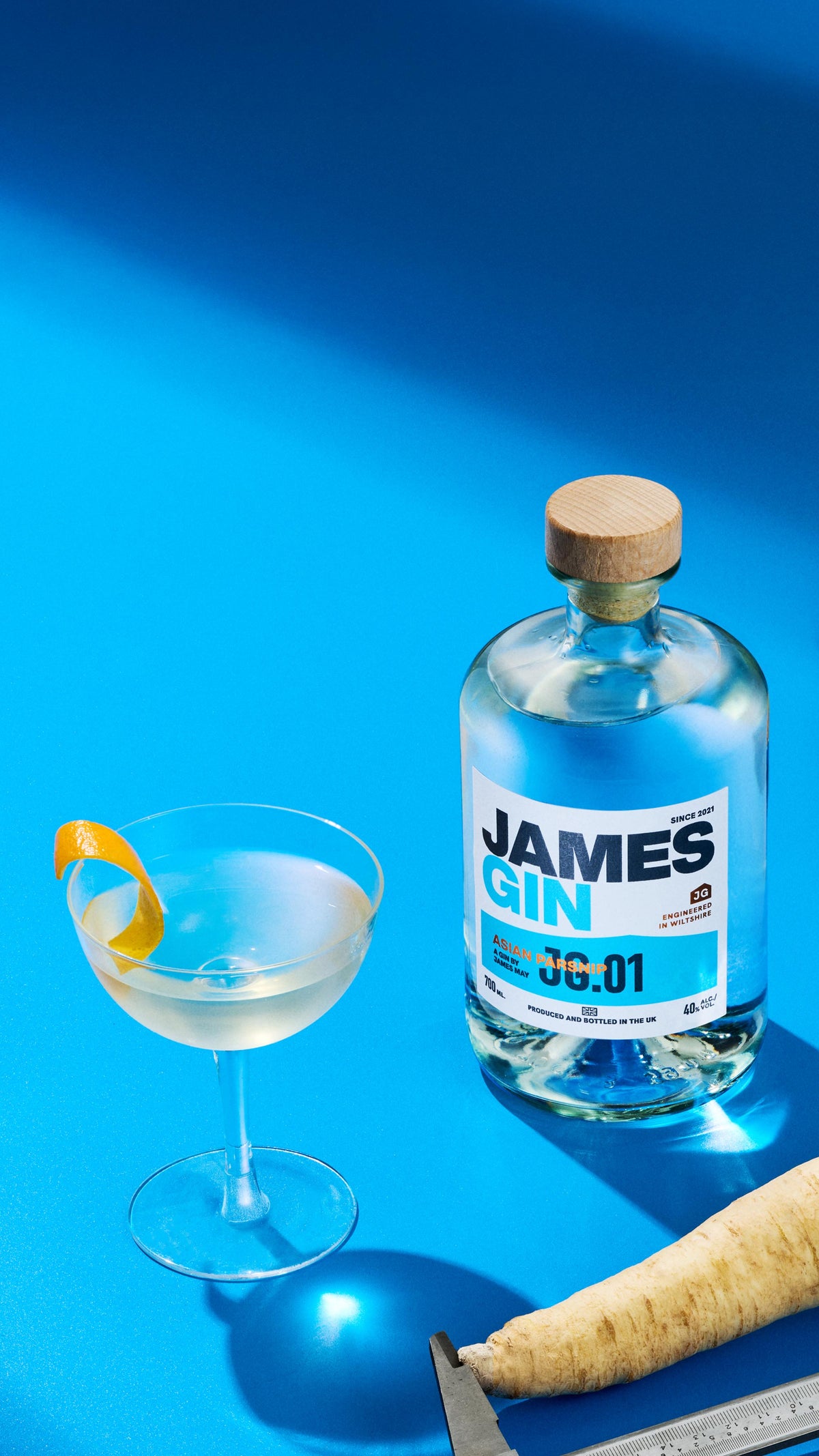 A James gin bottle of asian parsnip alongside a  cocktail. a parsnip being measured by a callipers, a bowl of juniper berries and a bowl of orange peel