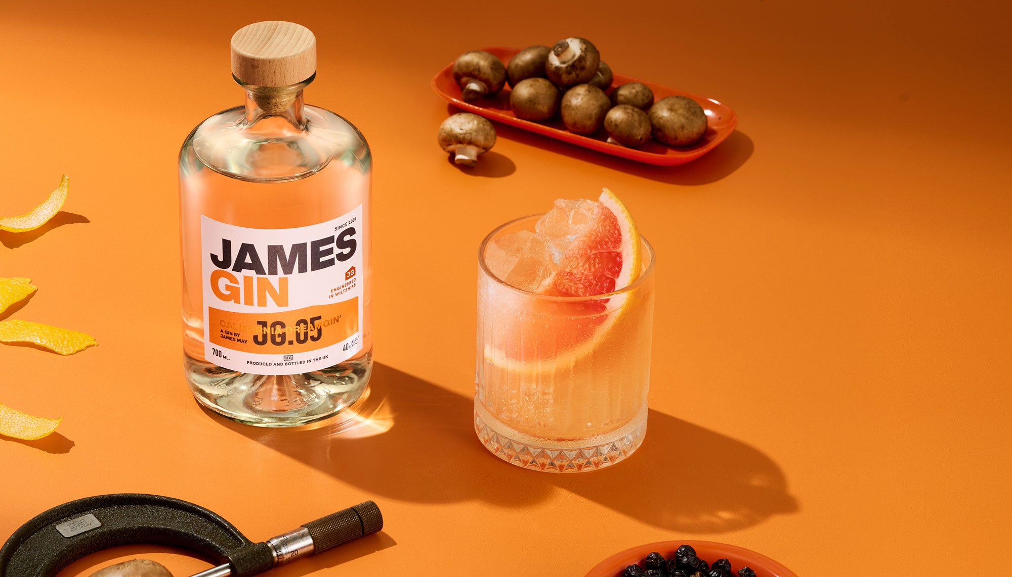 A James Gin bottle of California dreamgin' beside an orange cocktail with grapefruit slice, a plate of button mushrooms, a bowl of juniper berries, lemon peel and a callipers measuring a mushroom