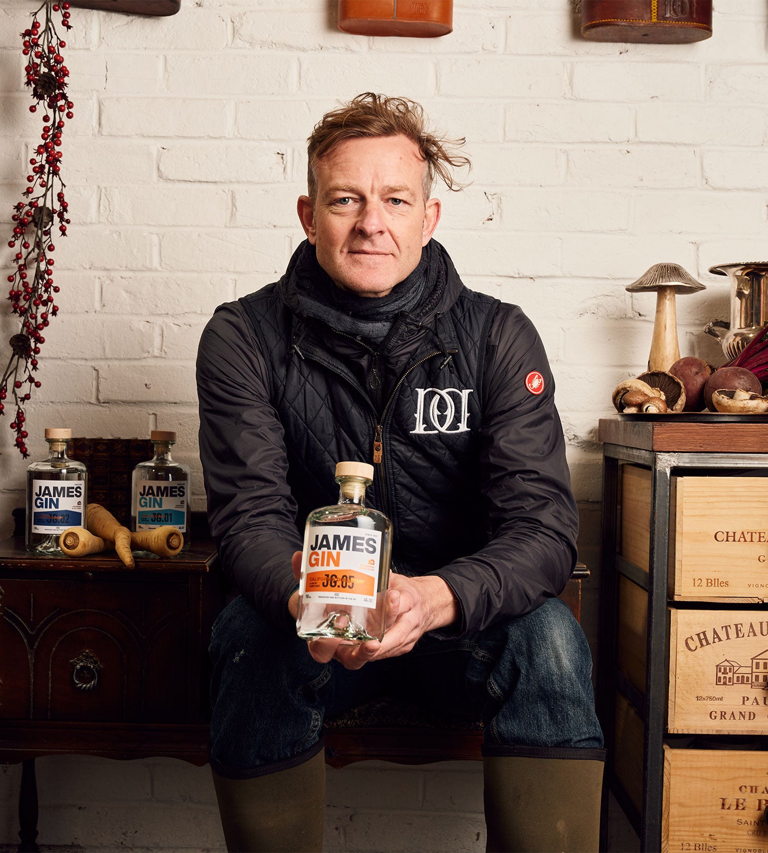 Hugh in the shed sitting down holding a bottle of california dreamgin' surrounded by other bottles of james gin and botanicals 